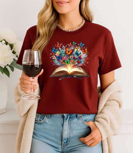 Person wearing a maroon t-shirt with a colorful book graphic, holding a glass of red wine.