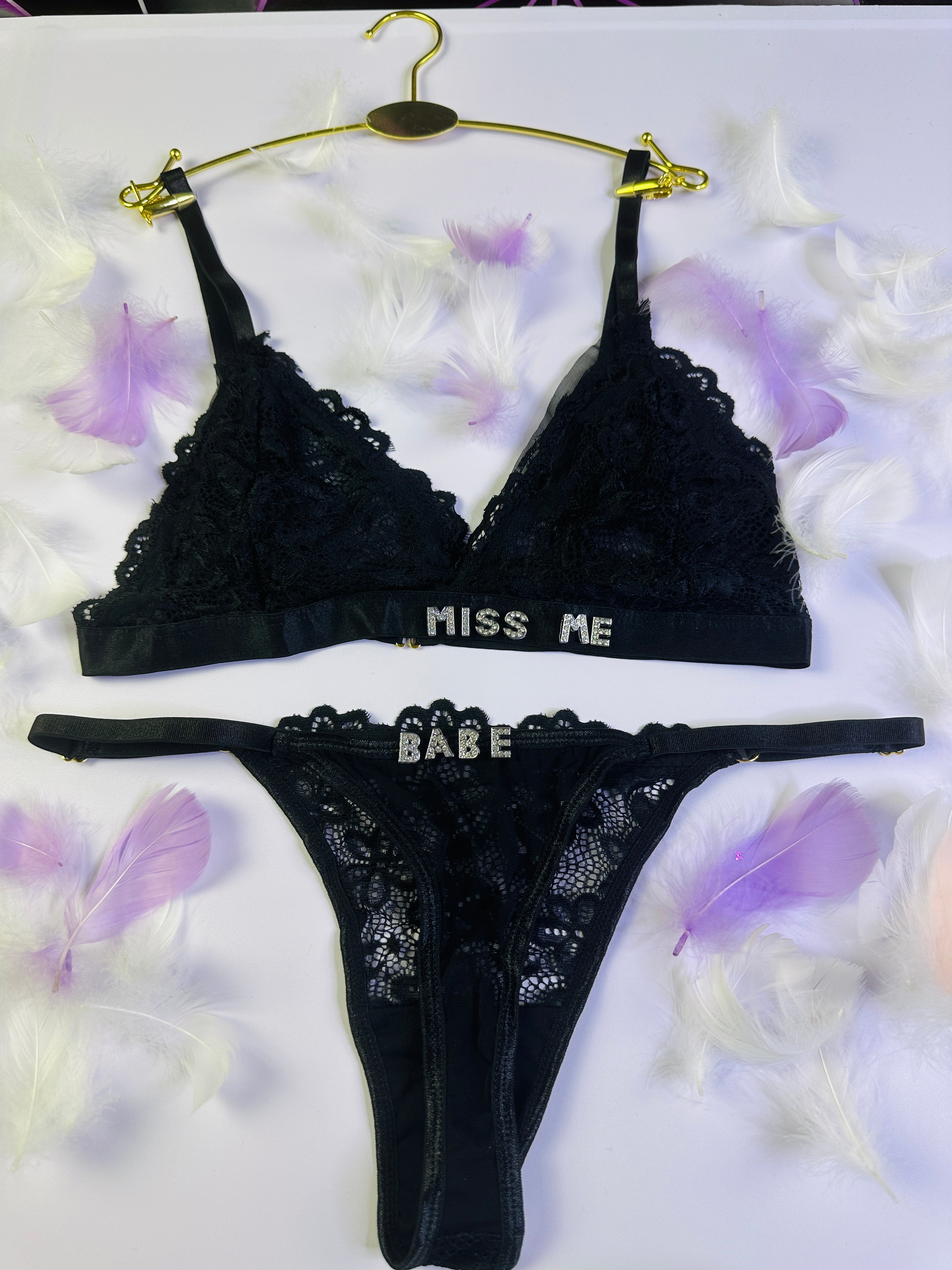 Black lace lingerie set with 'Miss Me' and 'Babe' text on a white background with purple flowers.